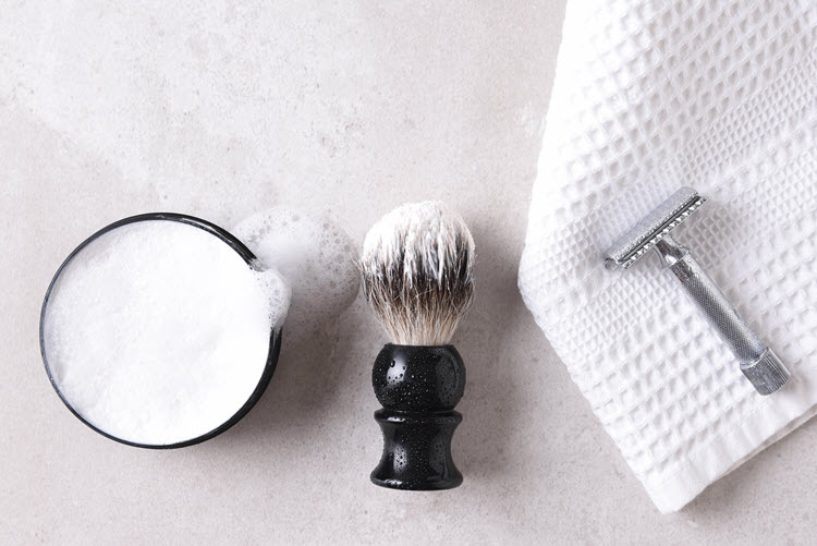 Safety razor on a towel with brush and soap on a gray tile surface.
